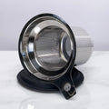 Stainless steel one-cup loose leaf tea infuser resting on its silicone lid coaster beside a ceramic mug
