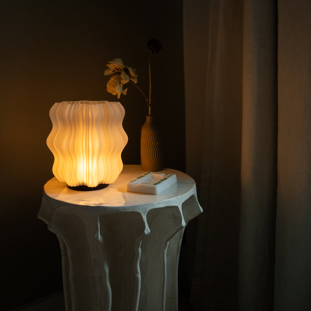 Cordless mini wavy sculptural table lamp with warm glowing filament on a nightstand